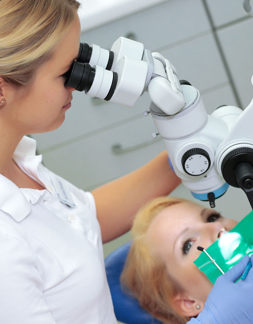 Eine Zahnärztin führt eine präzise zahnmedizinische Behandlung mit einem Dentalmikroskop an einer Patientin in einer modernen Zahnarztpraxis durch.