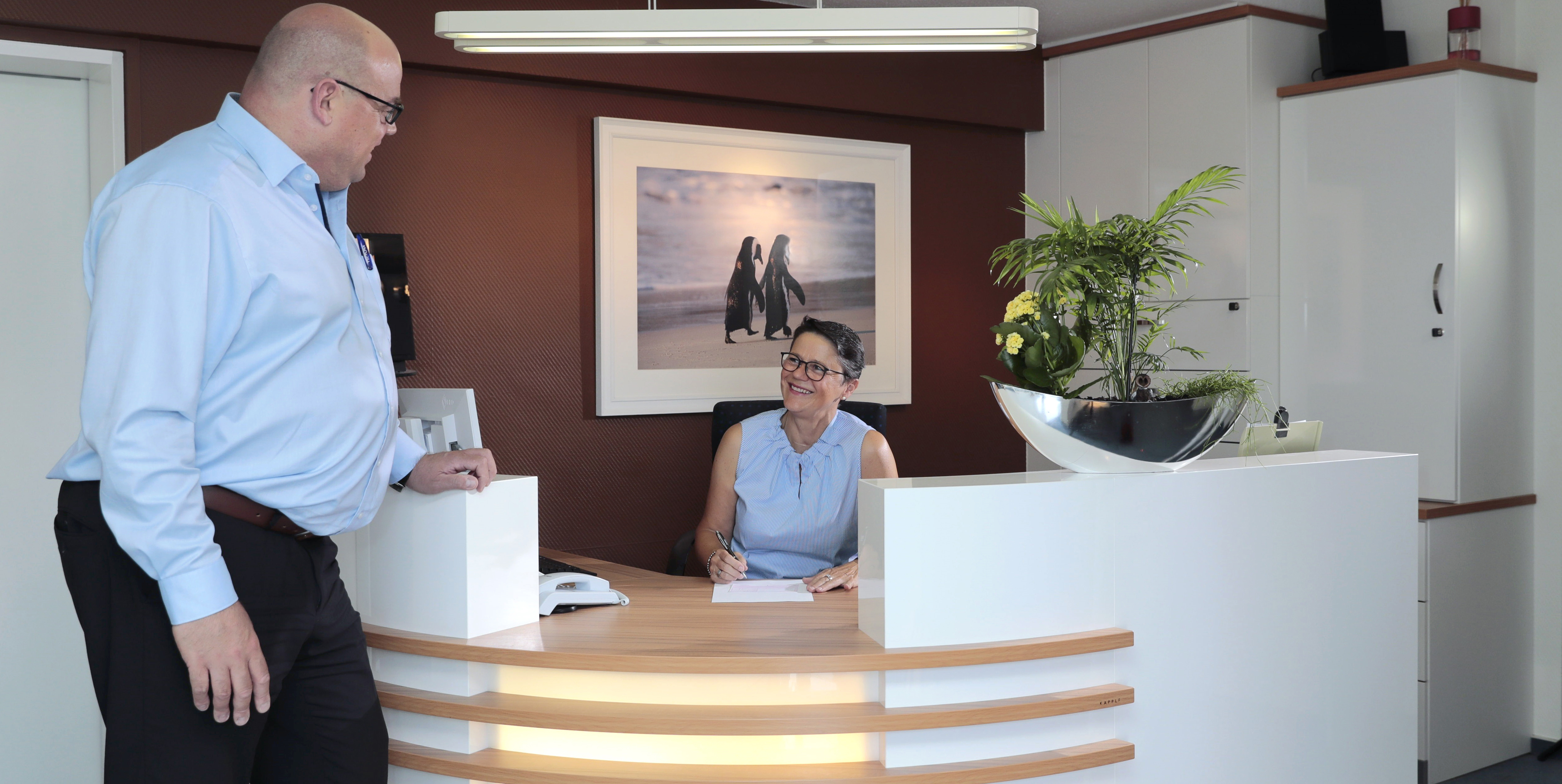 Rezeption in der Zahnarztpraxis Thiele & Menk in Weilburg: Ein Patient steht an der Empfangstheke, während die Rezeptionistin freundlich mit ihm interagiert – ein Bild von der persönlichen und professionellen Atmosphäre der modernen Zahnmedizinpraxis.